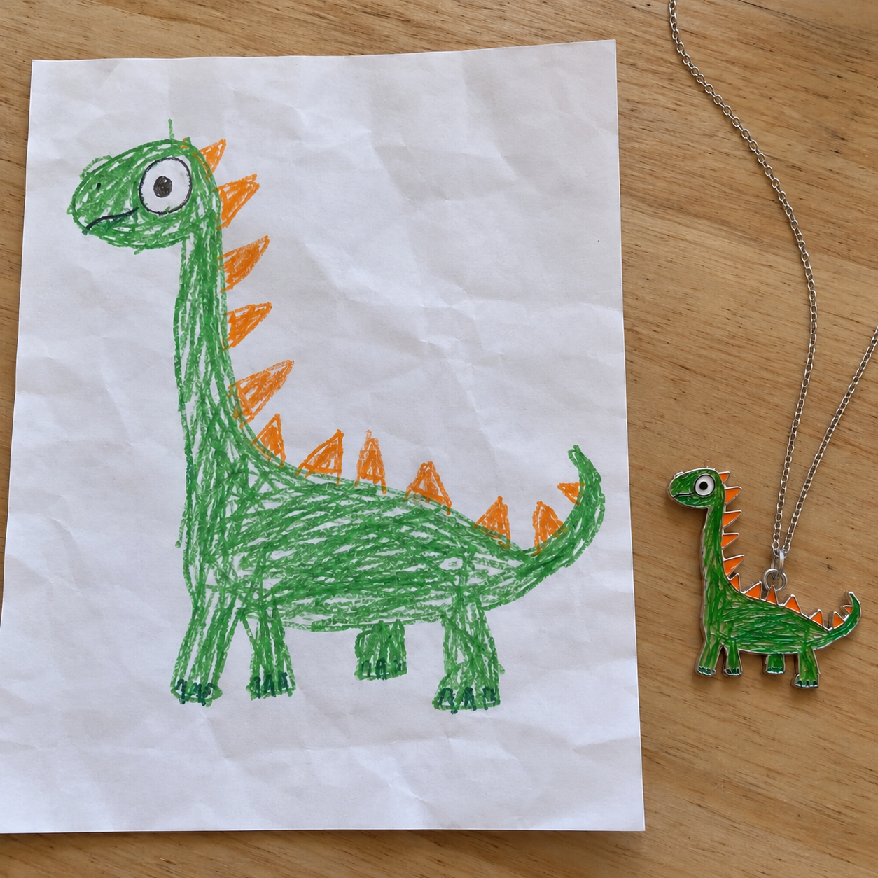 A child's green dinosaur drawing beside the matching Forever Color full-colour necklace