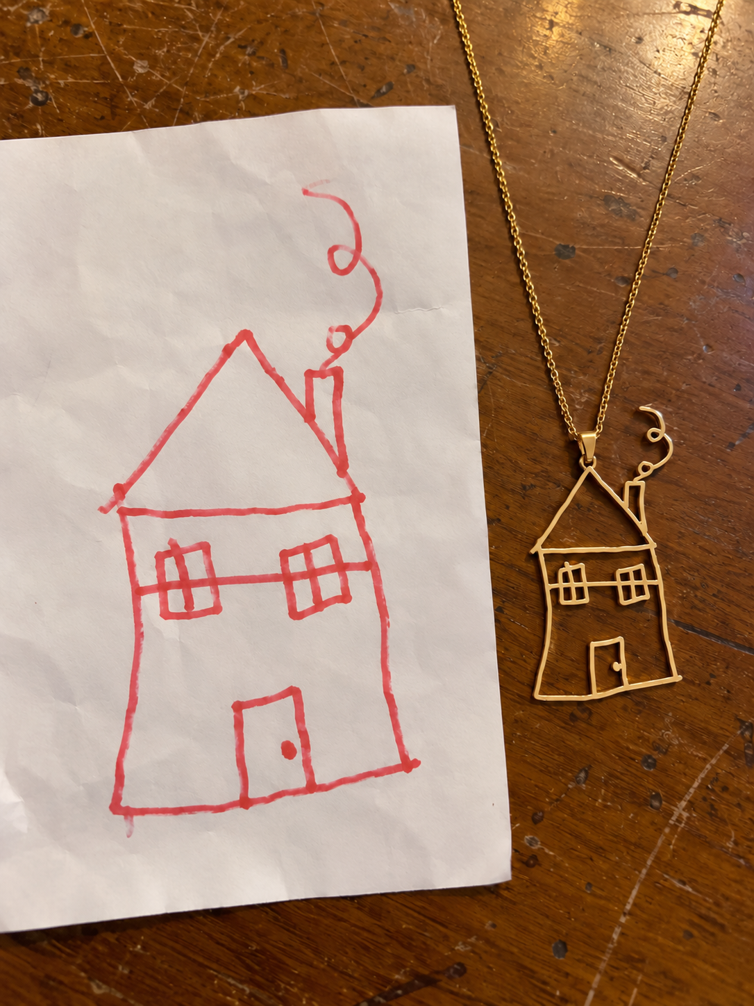 A child's red marker drawing of a house beside the matching gold cut-out house necklace